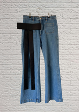 Load image into Gallery viewer, Light blue denim flare jeans with a structured black silk faille fabric bow pinned on patch pocket, hanging on a wire hanger against a white brick wall.