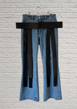 Load image into Gallery viewer, Light blue denim flare jeans with oversized blacksilk faille bow brooches pinned to pockets hanging from a metal hanger against a white brick wall.
